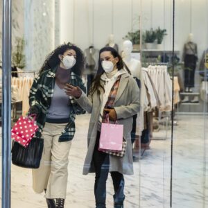 Full body of female friends in protective masks with shopping bags walking through glass doors of clothing store